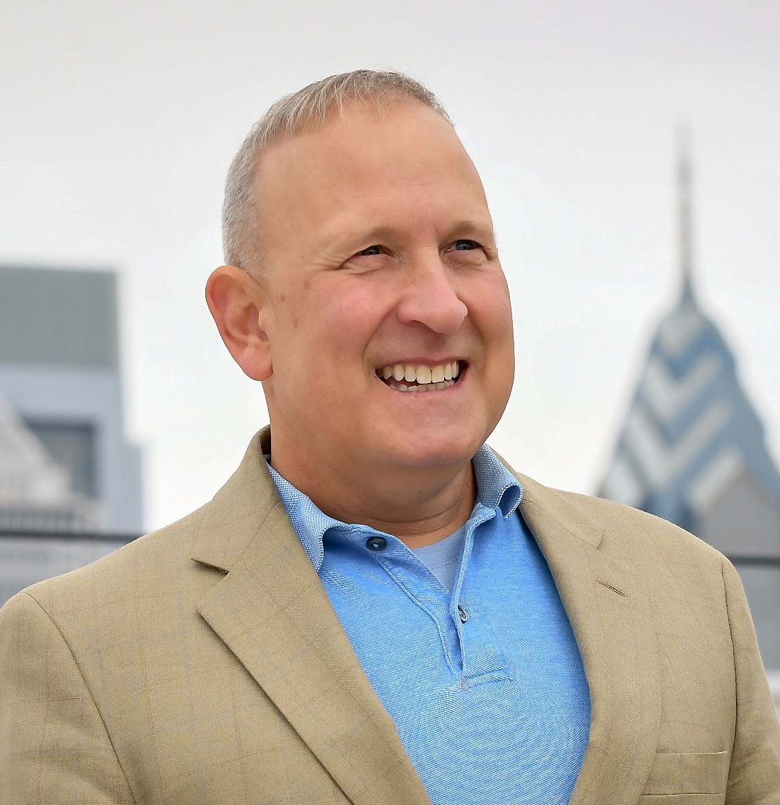 R Perry Monastero shown from the chest up standing with the Philadelphia skyline in the background. Perry is wearing a light blue polo shirt under a tan buttoned suit jacket. 