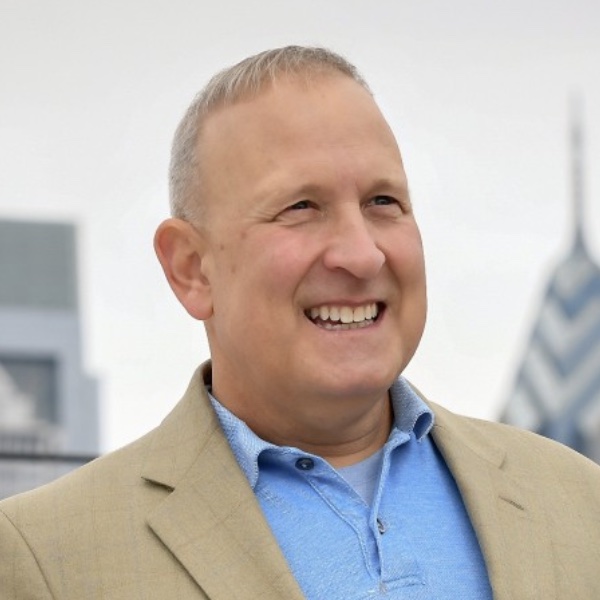 Photo of R Perry Monastero smiling with the Philadelphia skyline in the background. Perry is wearing a tan suit jacket over a blue polo shirt. 
