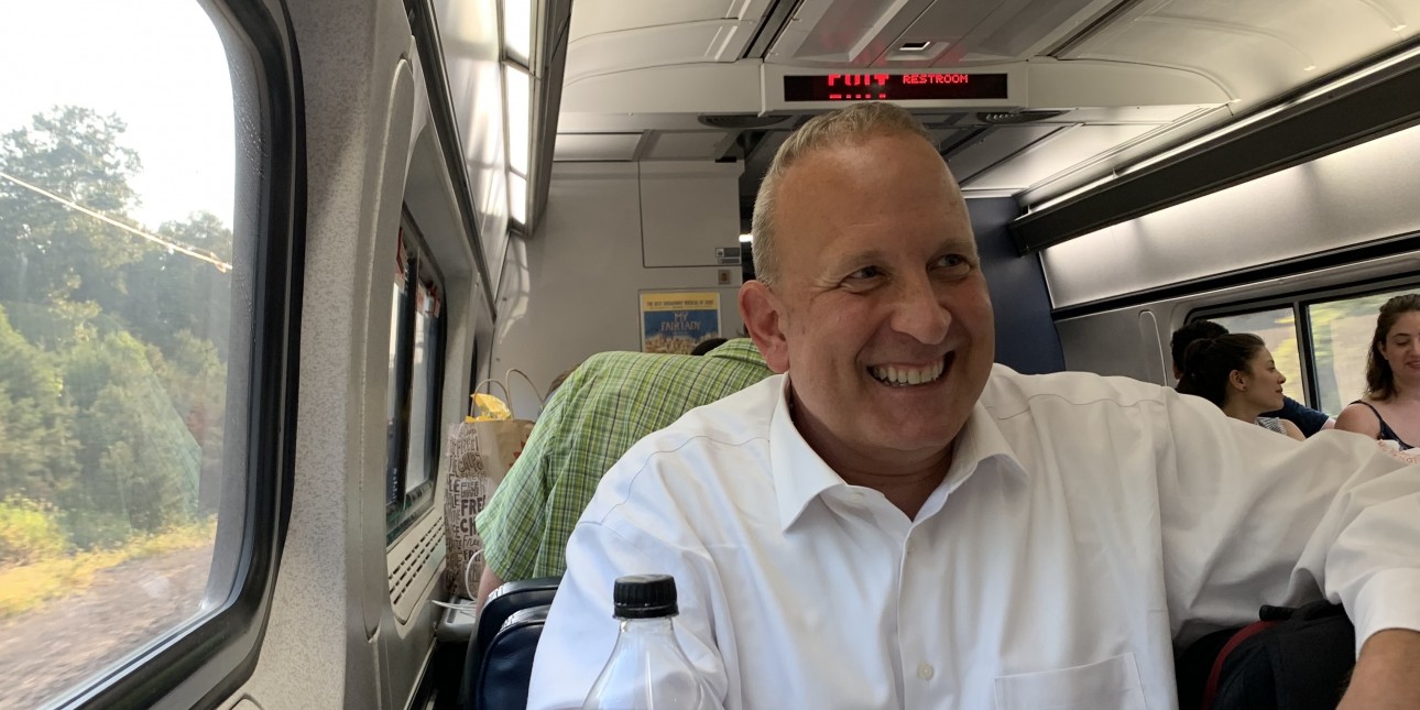 Perry wearing a white shirt smiling and sitting casually on an Amtrak train
